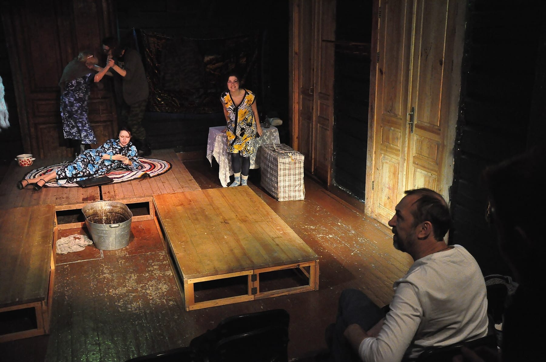 A director in a white T-shirt watches from the foreground as actors rehearse on a dimly lit wooden stage set