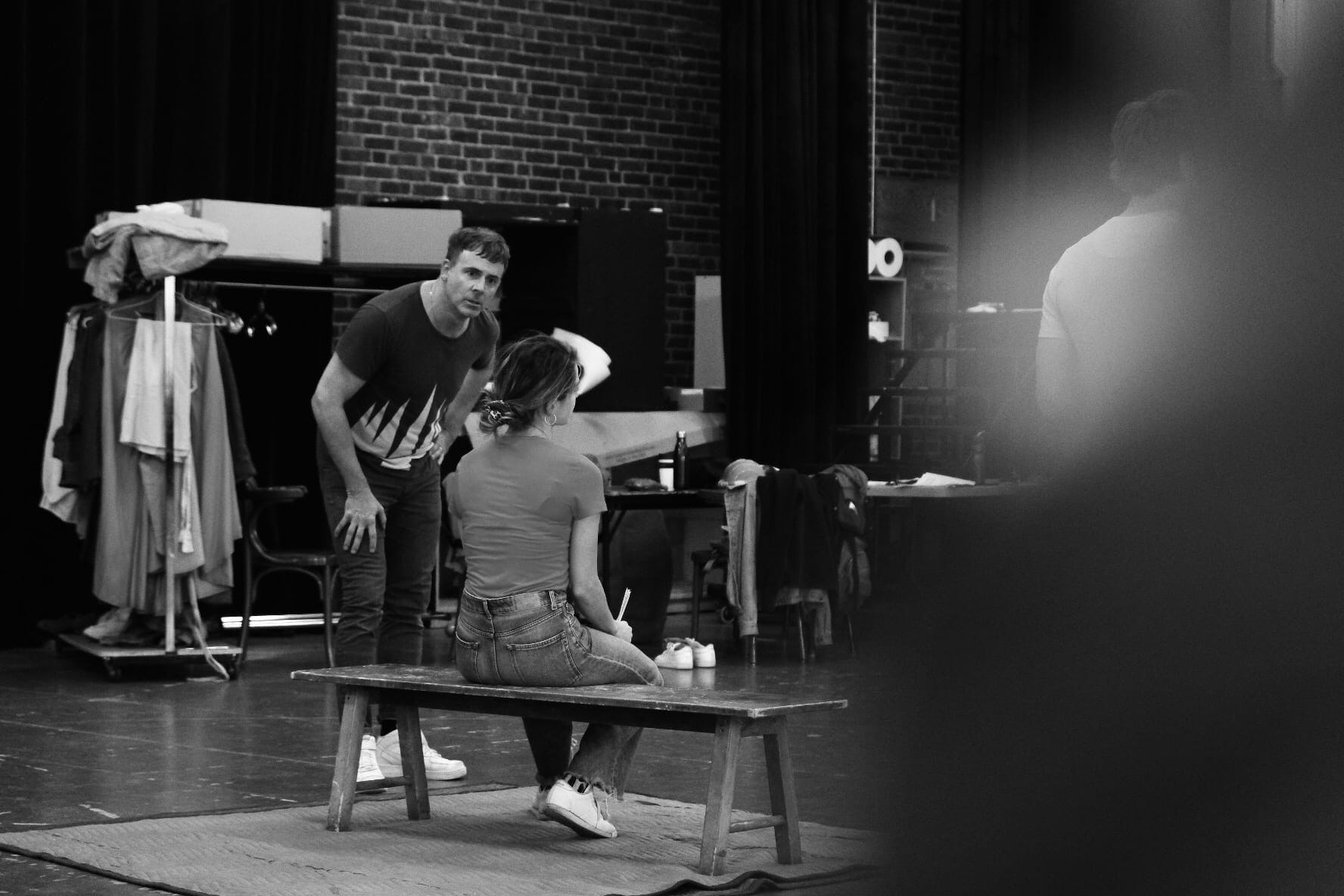 Two actors in a black-and-white photograph rehearsing a scene backstage, with a costume rail and brick wall behind them